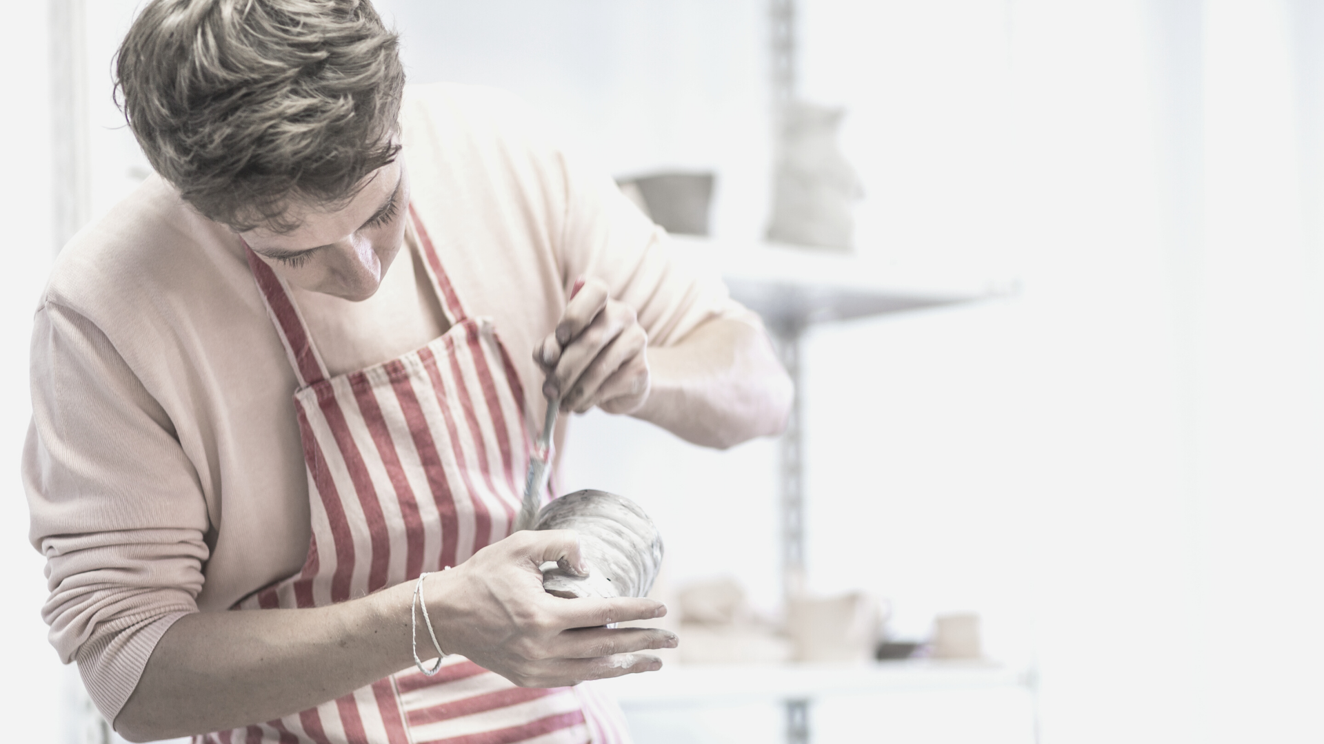 man in apron using a brush to glaze a small ceramic pot